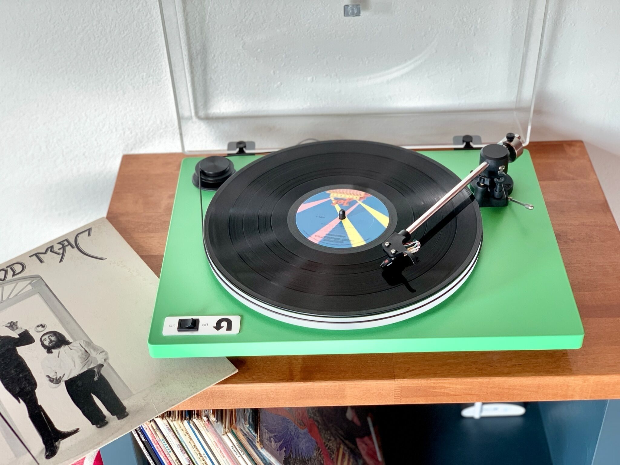 A turntable on a green stand with a vinyl record, a small shelf with a picture, and a row of books below.