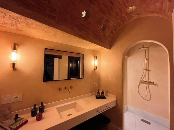 A bathroom with a large mirror, two wall-mounted lights, a shower, and a vanity with a sink and two bottles. - La Casa Juan Ranas (Granada)