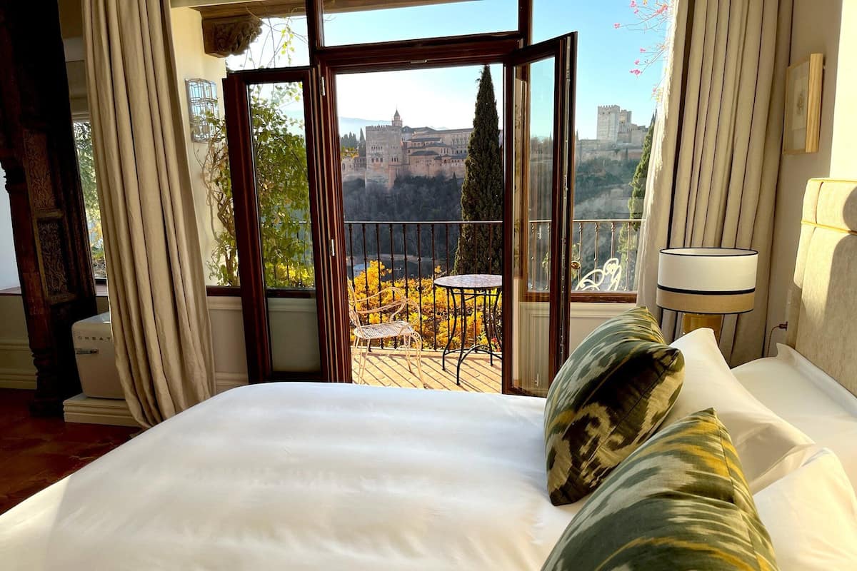 A hotel room with a large bed, a view of a castle through an open door, a balcony with a table and chairs, and a lamp on the side.