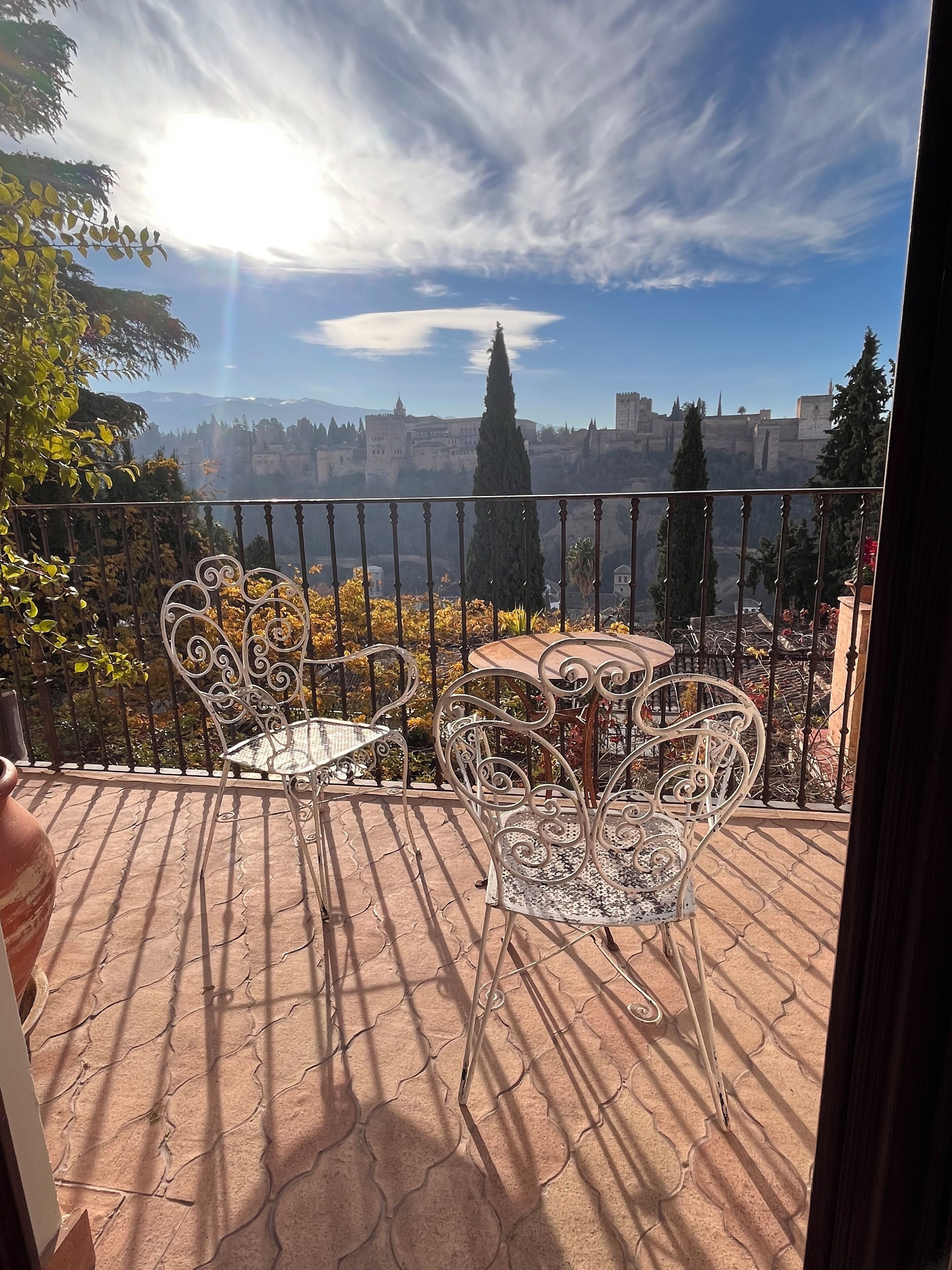 A balcony with a wrought iron railing, two metal chairs, and a small table. In the background, there is a view of a historic building with towers and a clear sky.