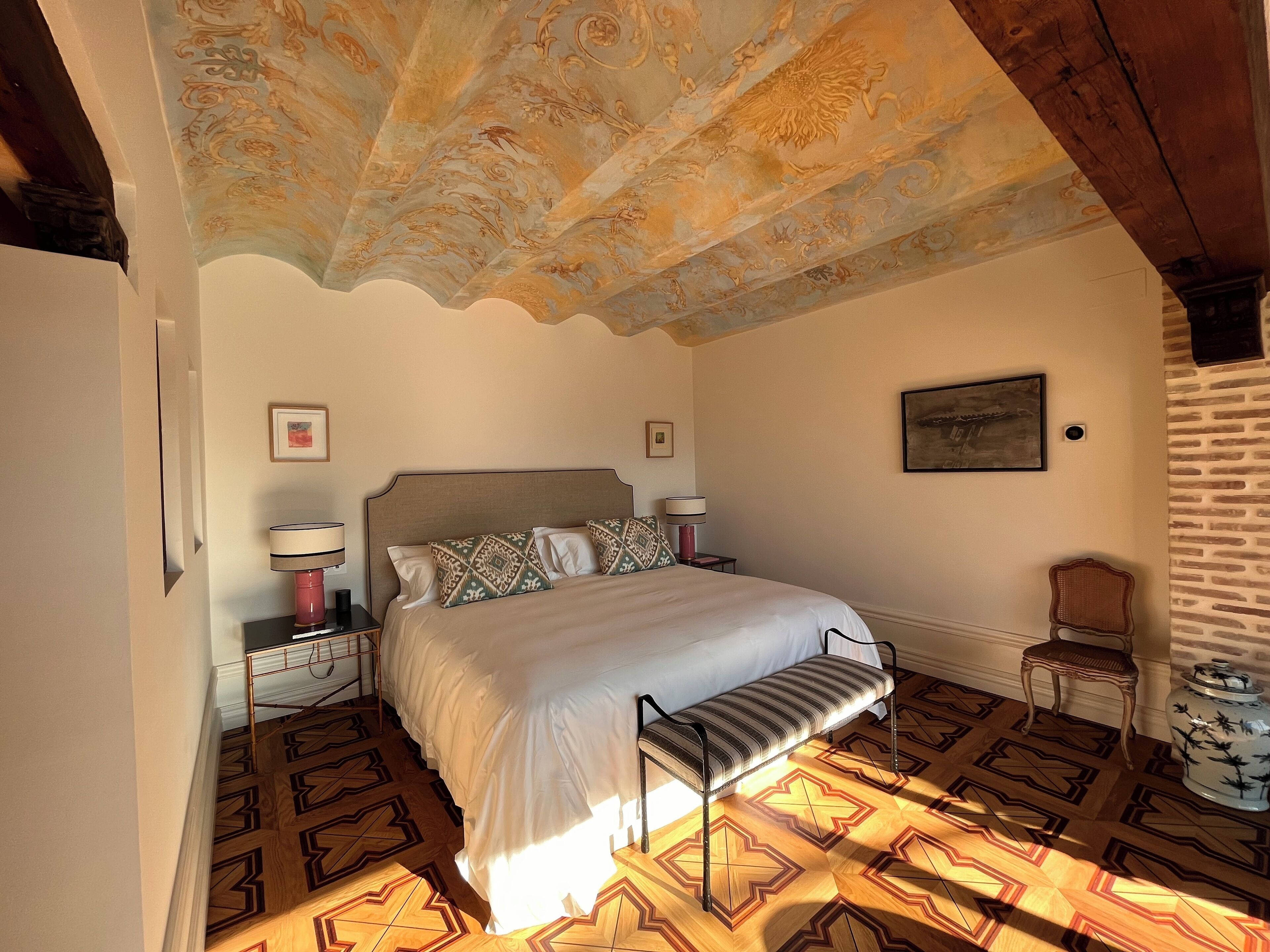 A bedroom with a bed, two bedside tables, a bench, a chair, and a decorative ceiling.