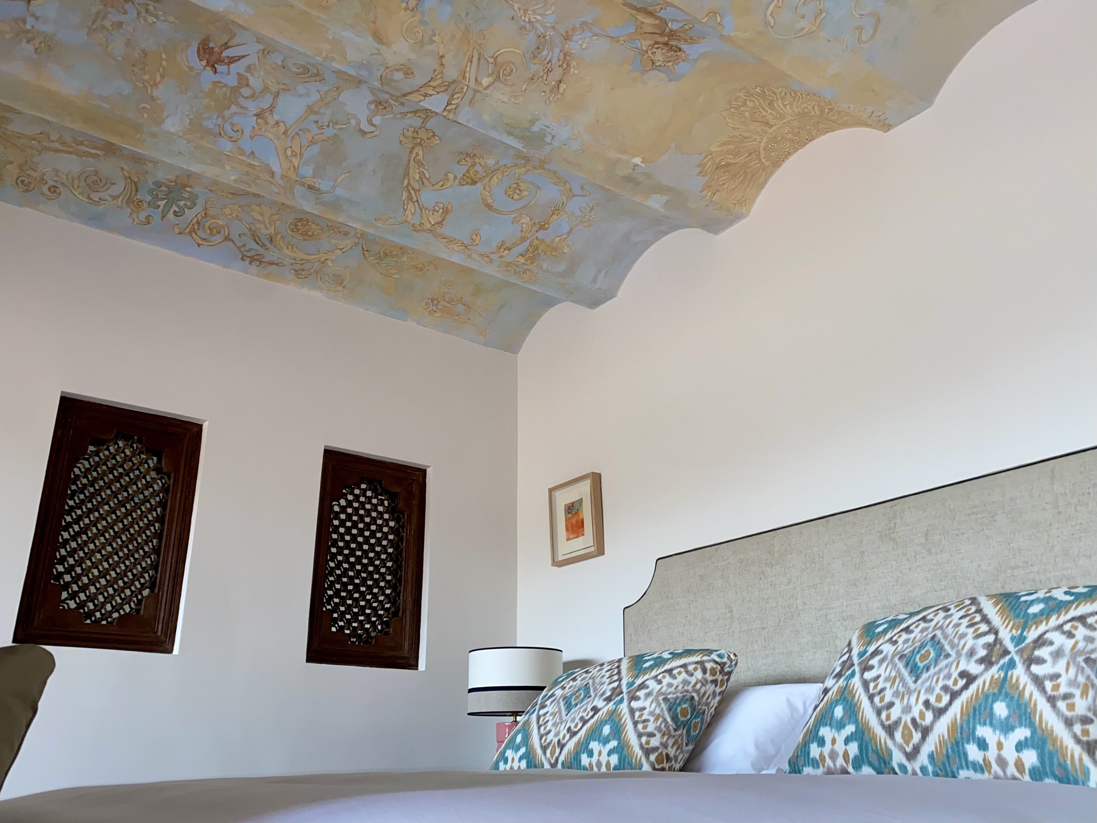 A bedroom with a patterned ceiling, a bed with patterned pillows, and two framed pictures on the wall.