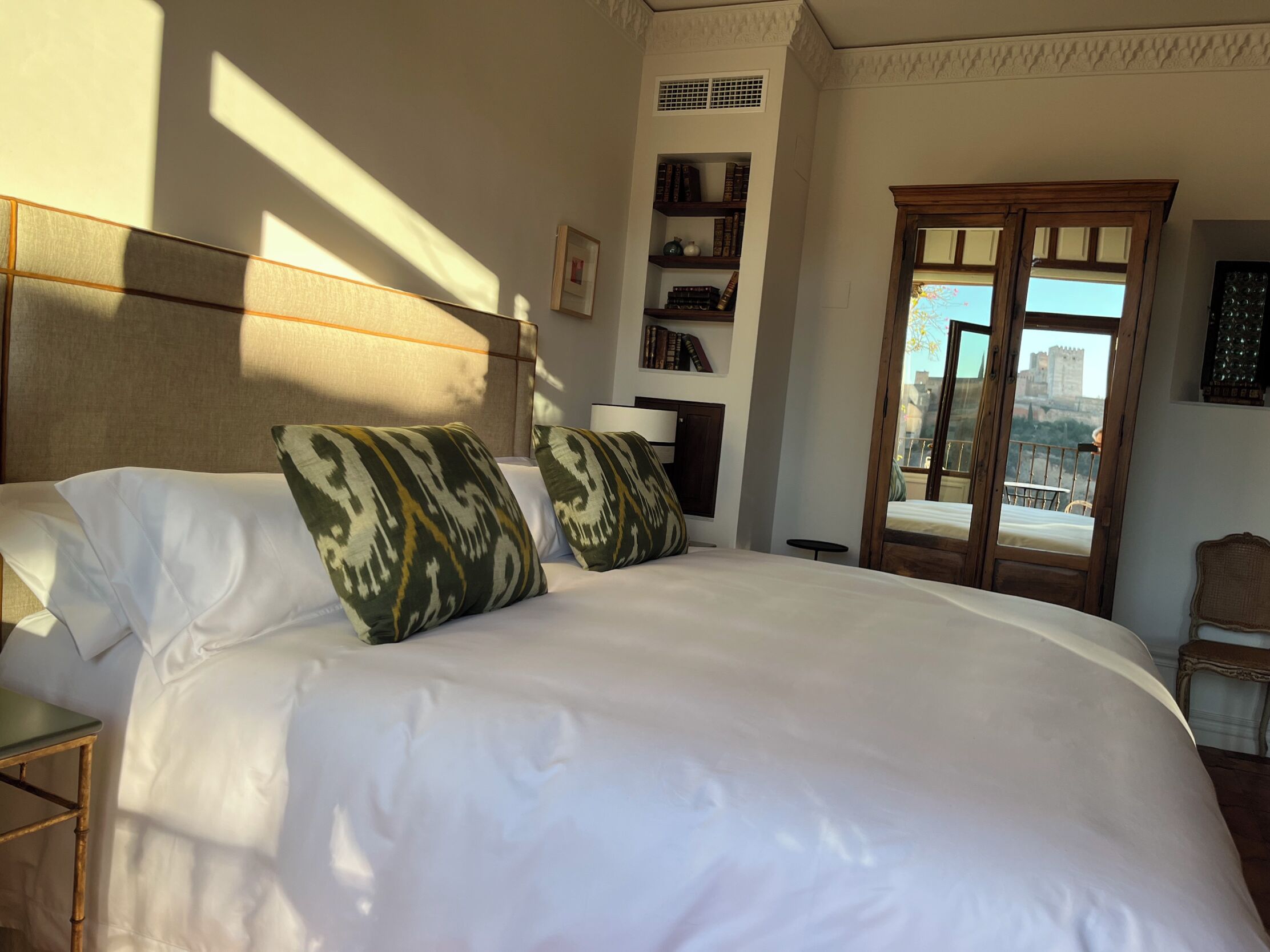 Une chambre d’hôtel avec un grand lit, une tête de lit, une fenêtre donnant sur un château et une bibliothèque remplie de livres.