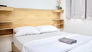A neatly made bed with white bedding and a folded towel, wooden headboard with built-in shelves, and a small potted plant on the shelf.