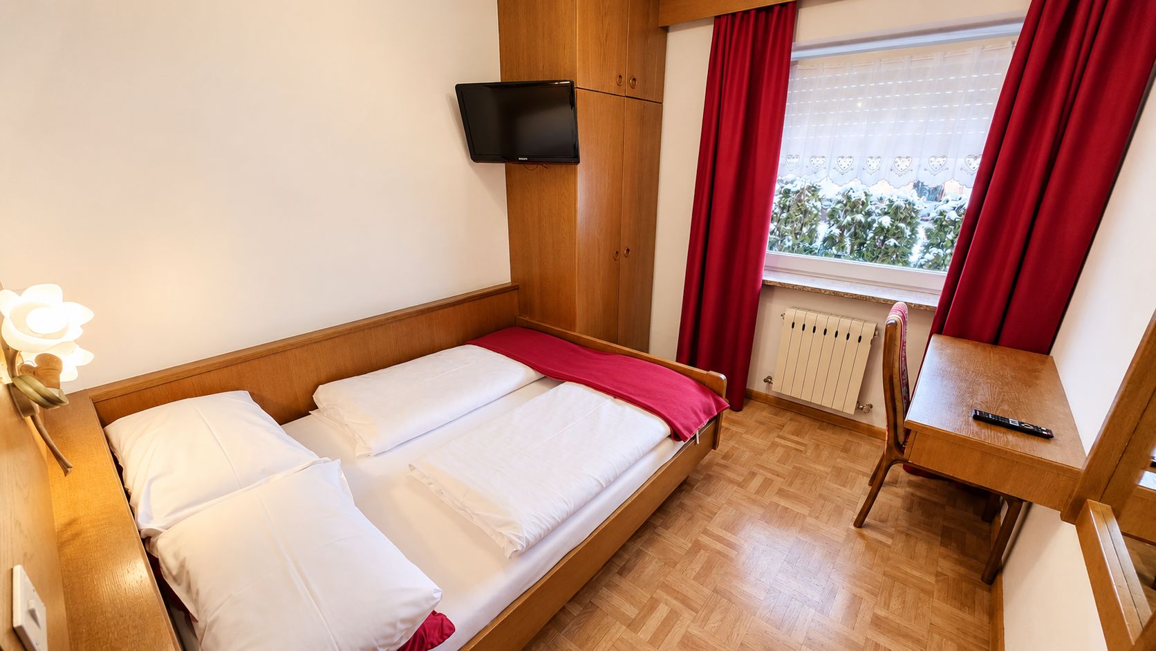 Una cama individual con ropa de cama blanca y una manta roja, una mesita de noche de madera con un control remoto, un televisor montado en la pared y una ventana con cortinas rojas.