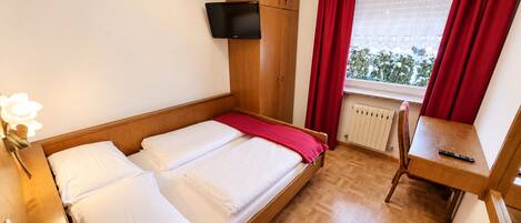 Una cama individual con ropa de cama blanca y una manta roja, una mesita de noche de madera con un control remoto, un televisor montado en la pared y una ventana con cortinas rojas.