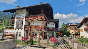 A traditional Alpine-style hotel with a balcony adorned with flowers, a sign indicating it is a restaurant, and a view of the surrounding mountainous landscape.