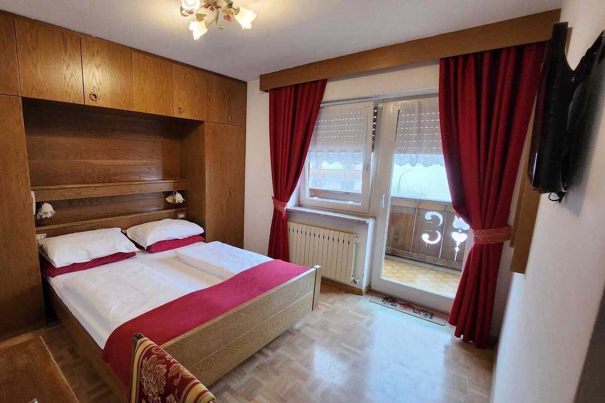 Ein Schlafzimmer mit Holz-Kopfteil, einem Bett mit weißer und roter Bettwäsche, einem Fenster mit roten Vorhängen und einem Heizkörper.