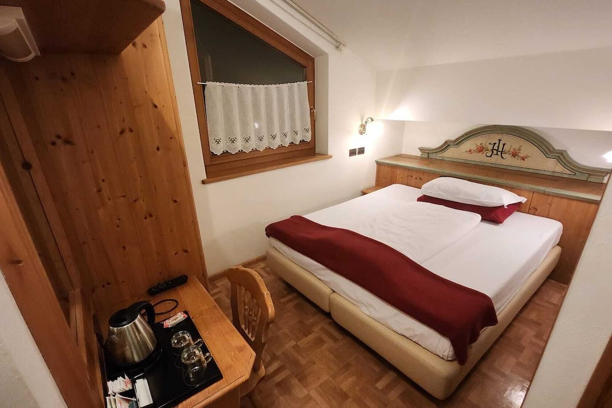 Ein kleiner, gemütlicher Raum mit einem hölzernen Bett, einem Nachttisch mit Wasserkocher und Tassen sowie einem Fenster mit weißem Vorhang.