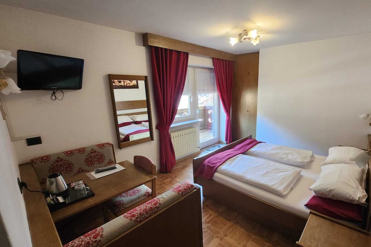 Ein kleines, gemütliches Zimmer mit Bett, Schreibtisch, Stuhl, Fernseher und einem fenster mit Vorhängen.