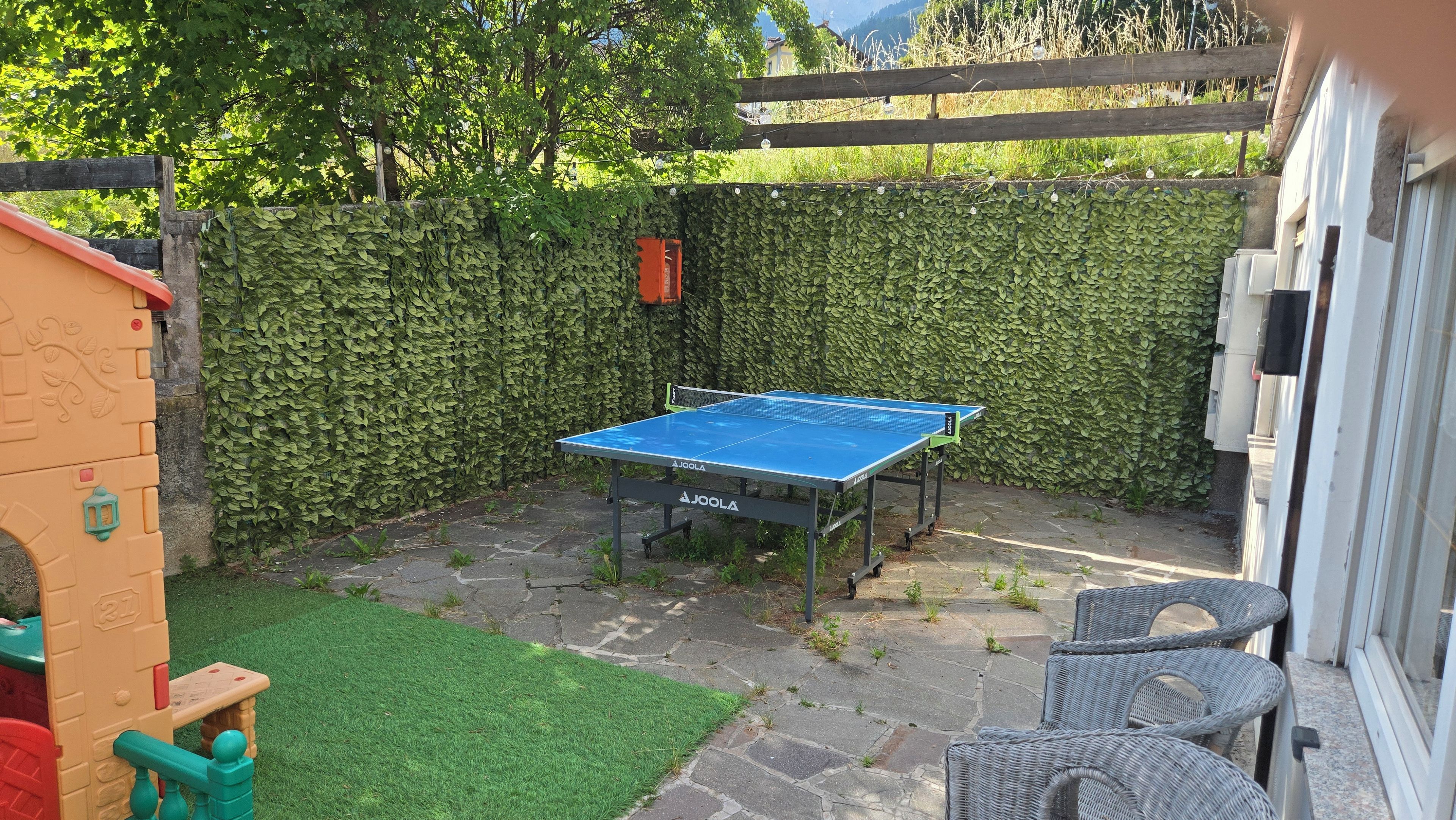 Una mesa de ping pong en un patio trasero con un seto verde, una estructura de juegos y mobiliario de mimbre.