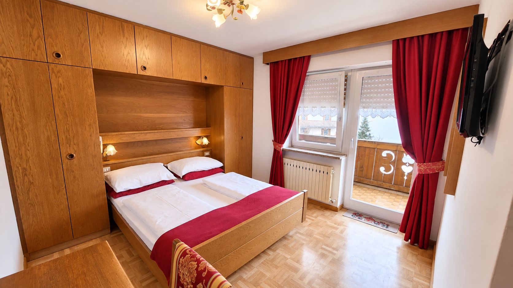 A bedroom with a wooden bed, a window with red curtains, and a view of a balcony.