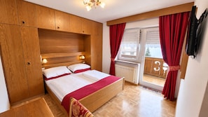 A bedroom with a wooden bed, a window with red curtains, and a view of a balcony. - InterHotel B&B (San Giovanni di Fassa)