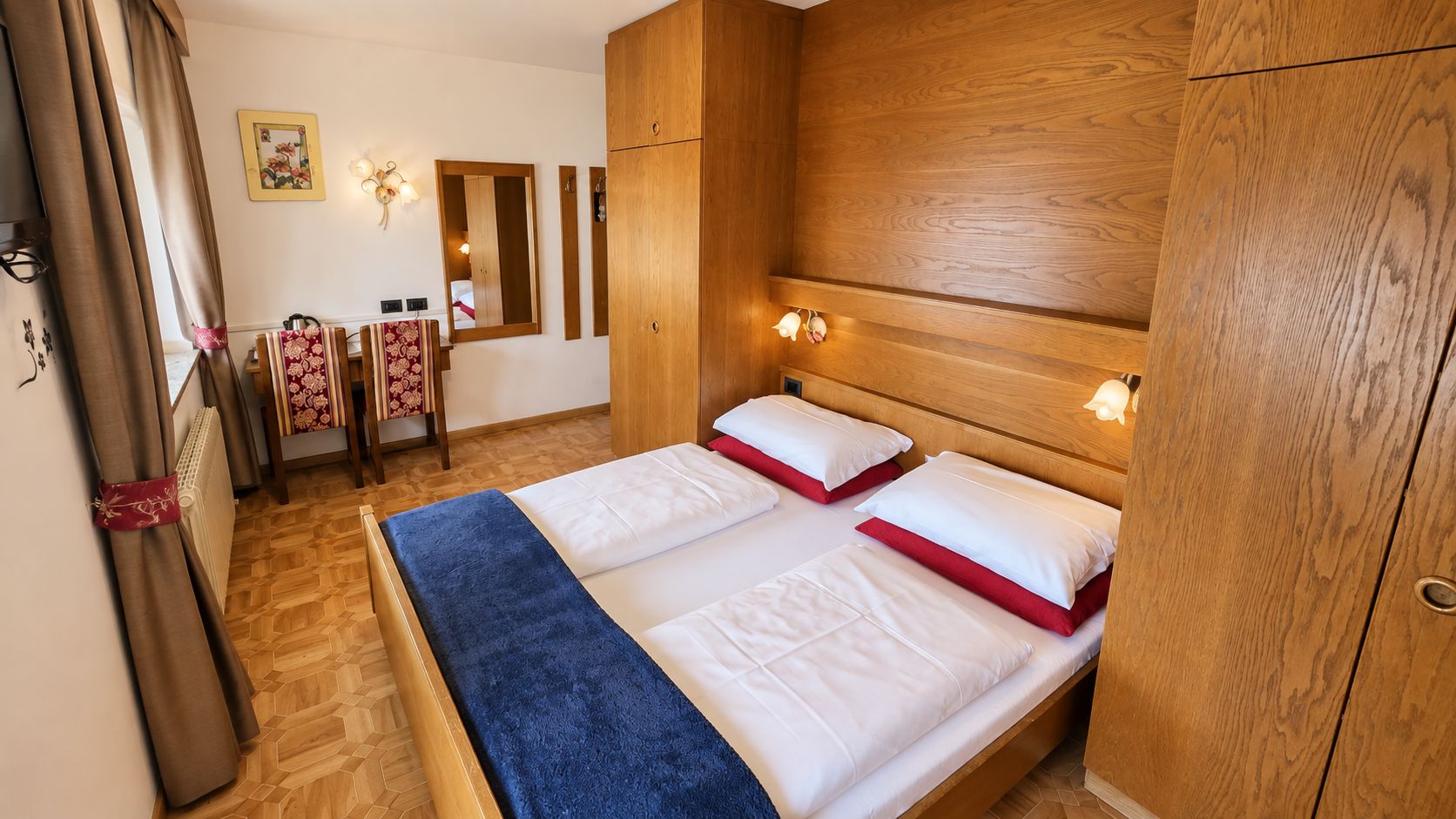 A hotel room with a wooden headboard, two beds with white bedding and red accents, a wooden floor, a mirror, and a radiator.
