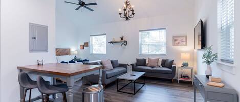 A spacious living room with a dining area, a ceiling fan, a chandelier, and large windows.
