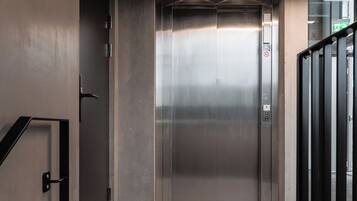 An elevator with a metallic interior, a handrail on the right, and a green exit sign above.