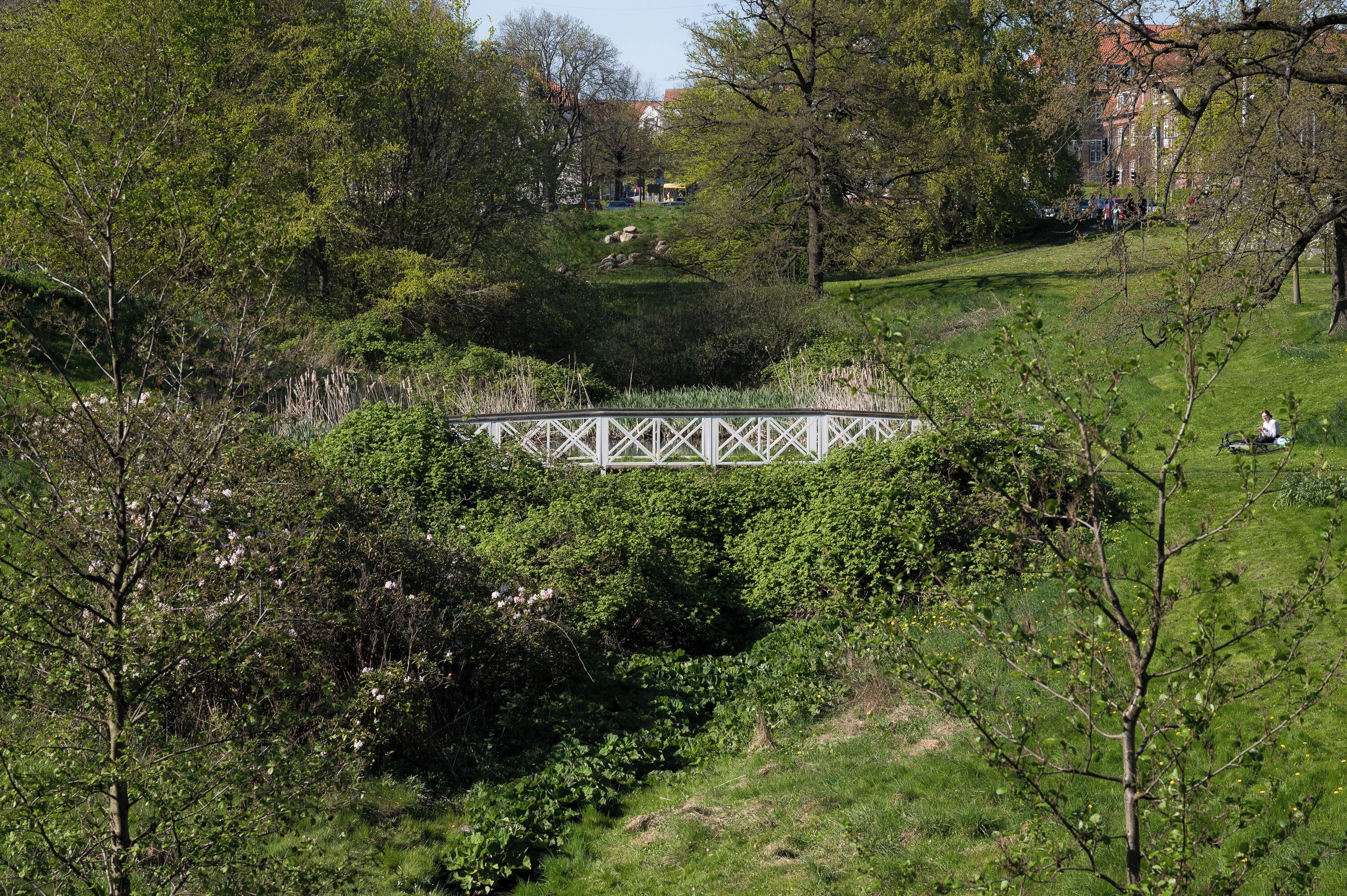 A park with a white bridge, lush greenery, and a distant building.