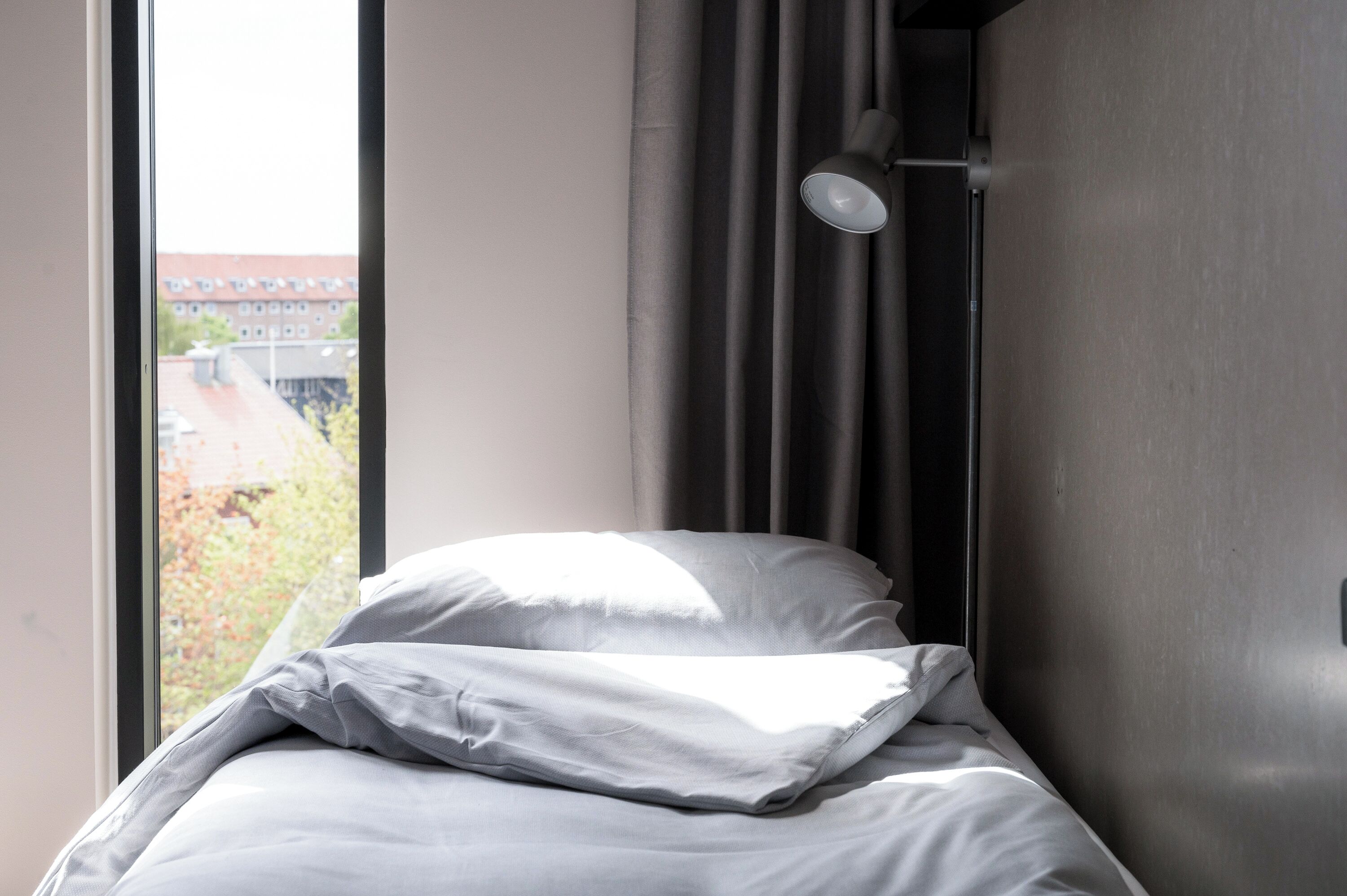 Una cama con ropa blanca, una ventana con cortinas y vistas a edificios.