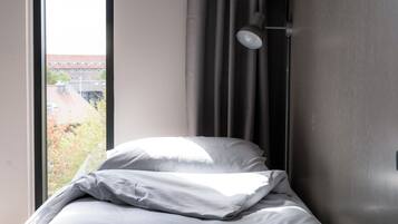 Una cama con ropa blanca, una ventana con cortinas y vistas a edificios.