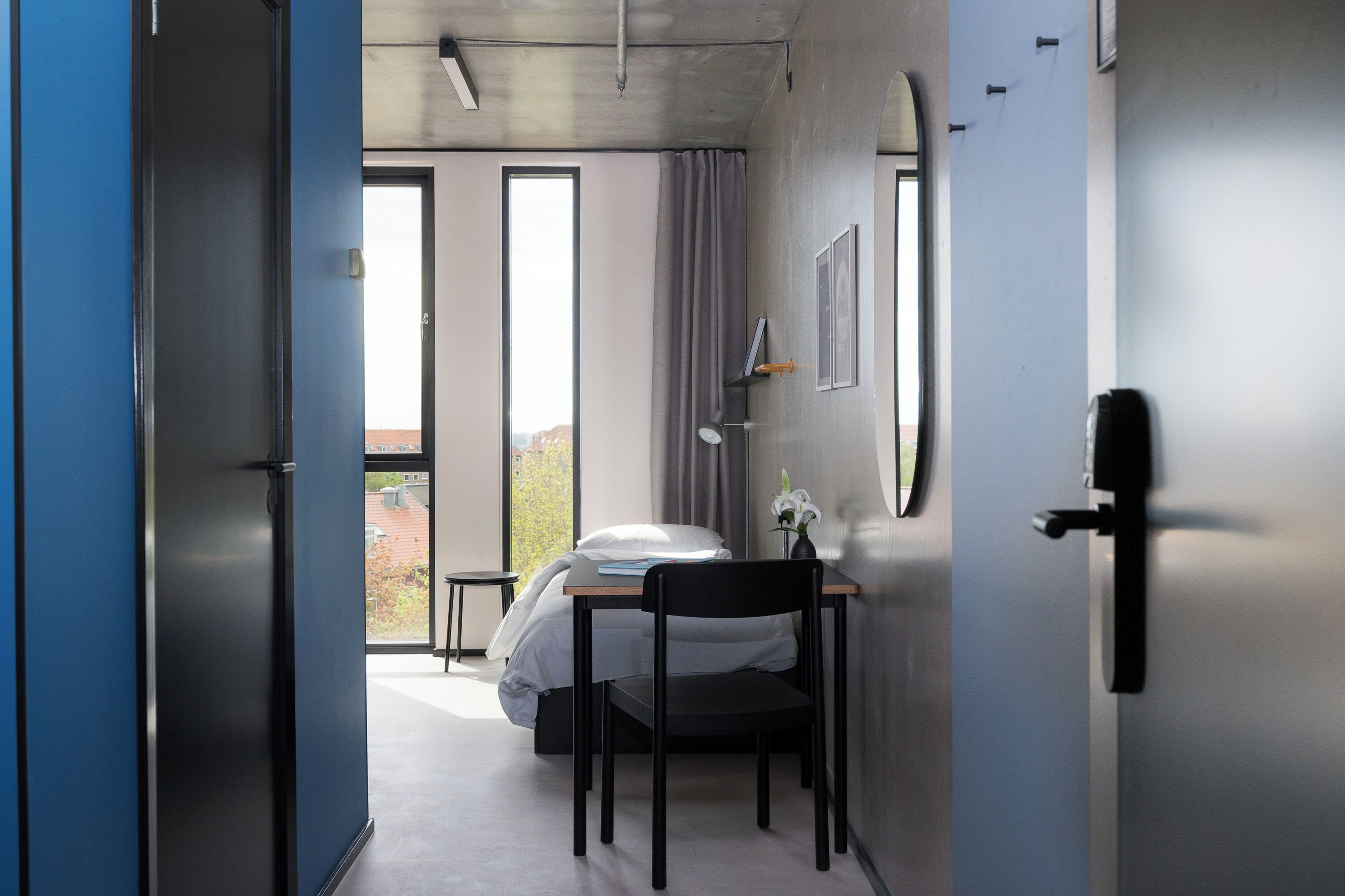 A modern hotel room with a bed, a desk, and a chair. There is a window with curtains, a door, and a wall-mounted light fixture.