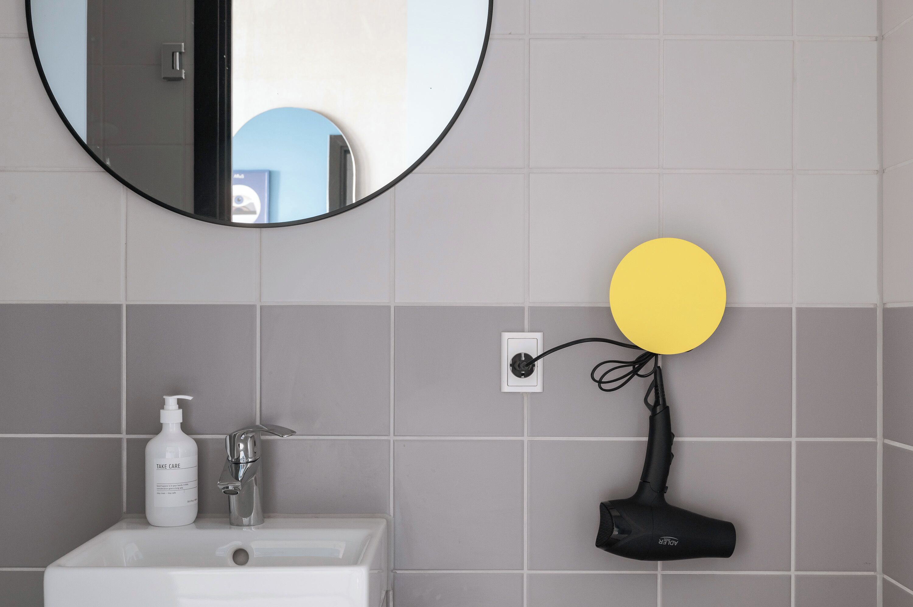 A bathroom with a sink, a mirror, a yellow light fixture, and a hairdryer on a wall.