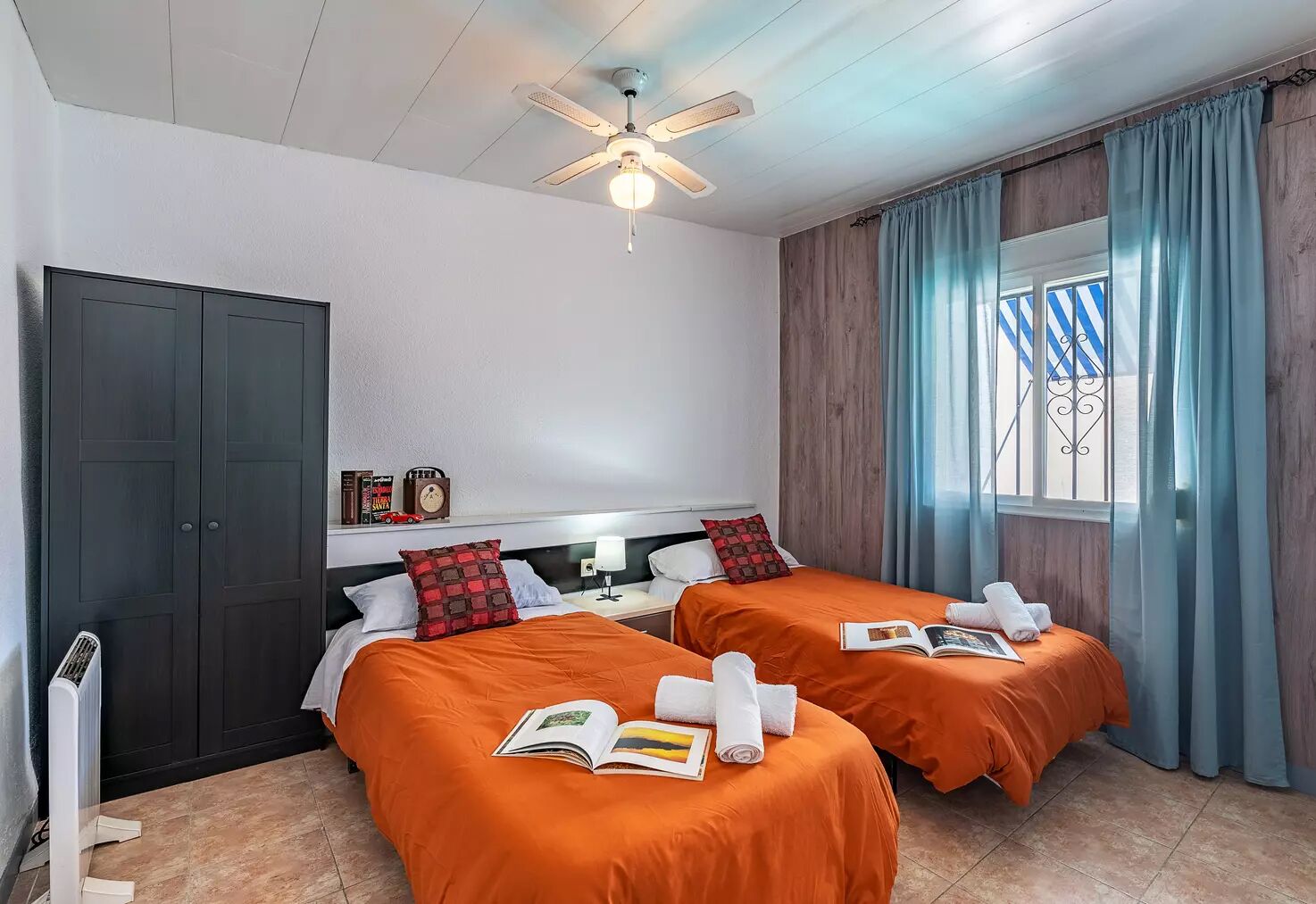A hotel room with two beds, a ceiling fan, a window with curtains, and a radiator.