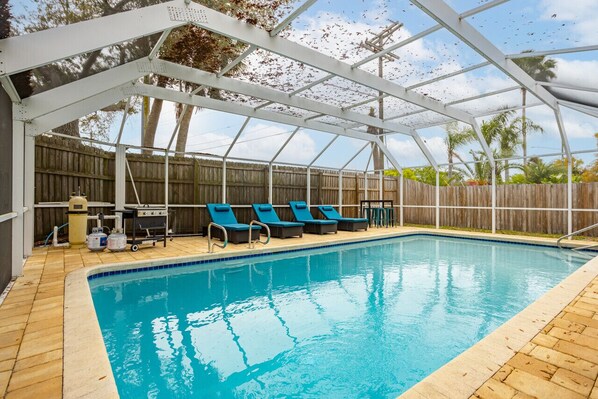A covered swimming pool with lounge chairs and a grill area.
