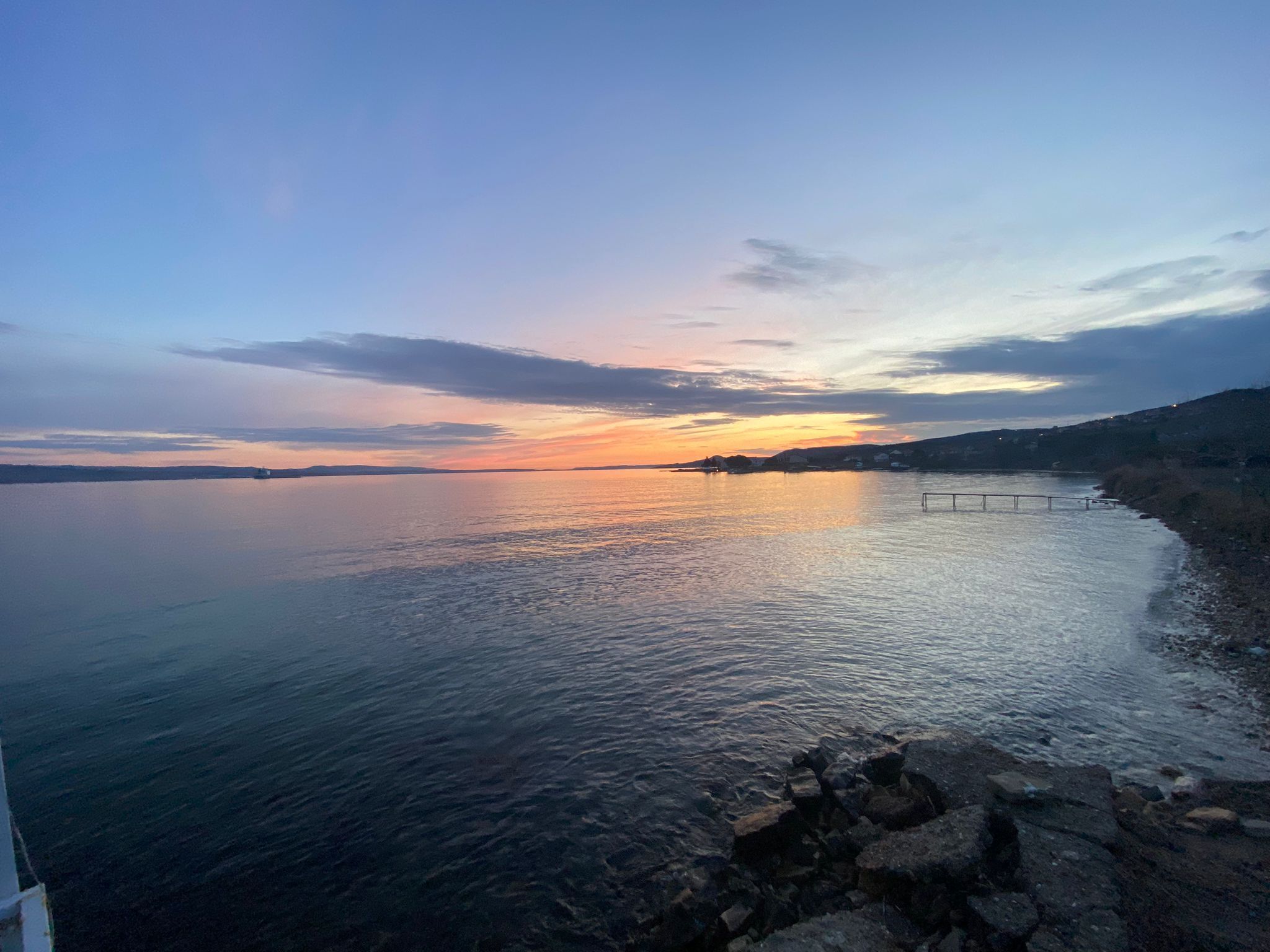 A calm lake at sunset with a rocky shoreline and a distant view of a small island.