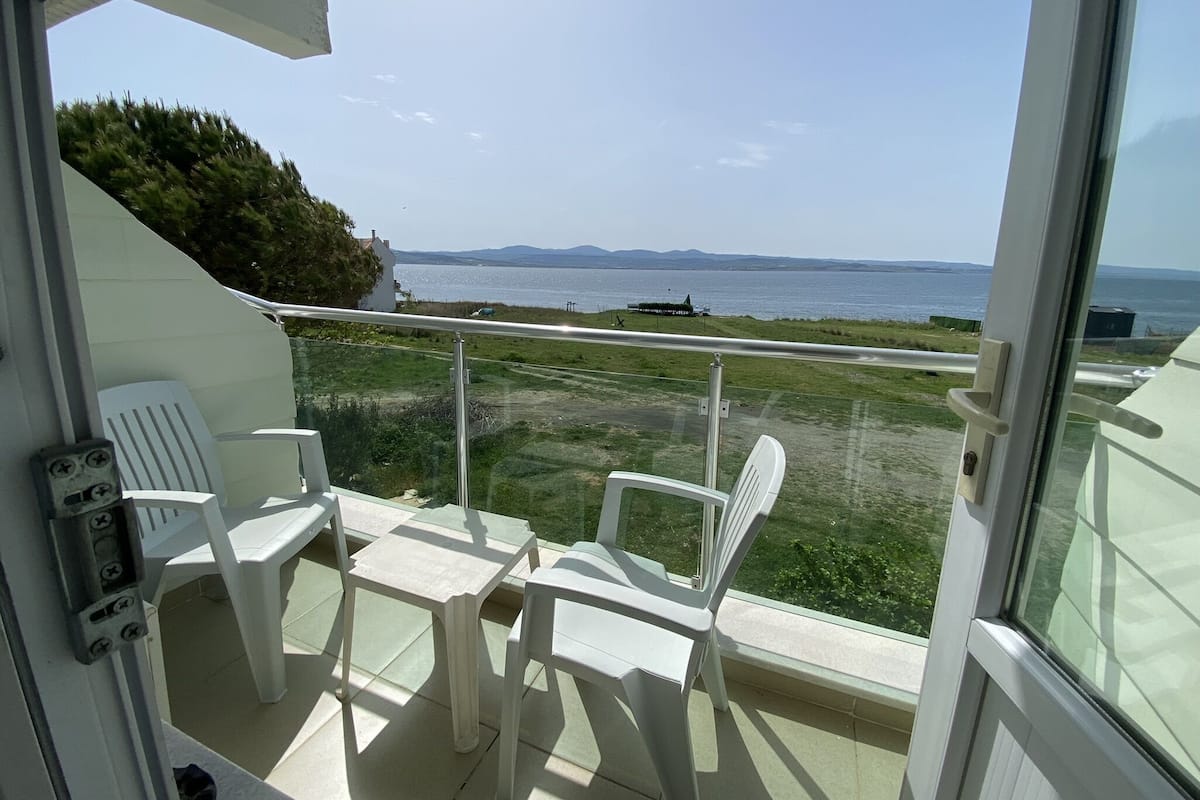 A balcony with two white plastic chairs and a small table, overlooking a grassy area and a body of water with distant landforms.