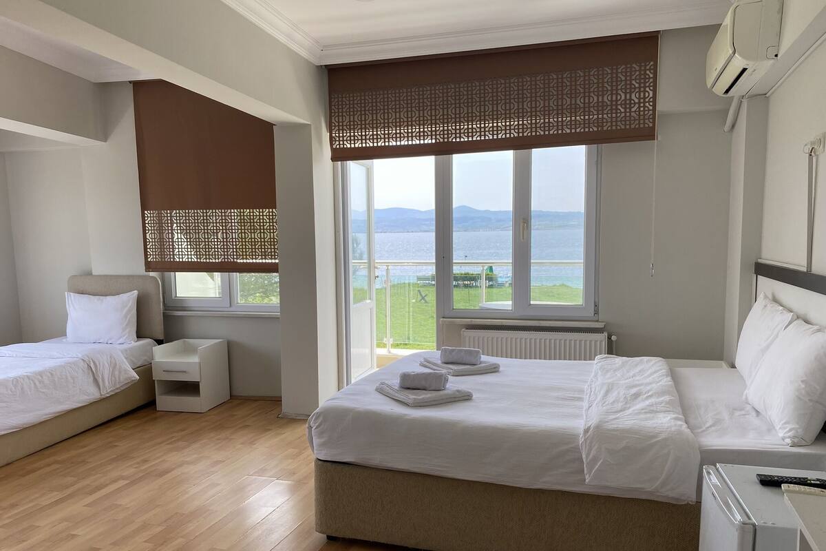 A hotel room with a large window offering a view of the sea, a bed with white linens, a bedside table, and a small seating area.