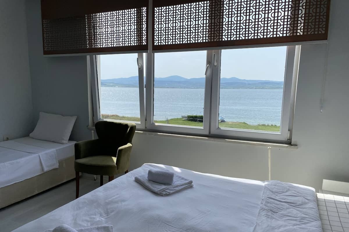 A room with two beds, a chair, and a large window with a view of the sea and mountains.