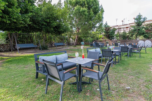 An outdoor seating area with tables and chairs, surrounded by trees and a stone wall.