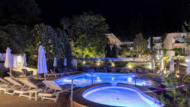 Un área de piscina iluminada de noche, con sillones reclinables y sombrillas, rodeada de árboles y edificios.