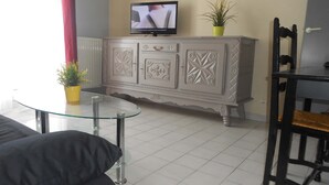 Un salon avec une table basse en verre, un canapé gris, un téléviseur posé sur un meuble et une petite plante.