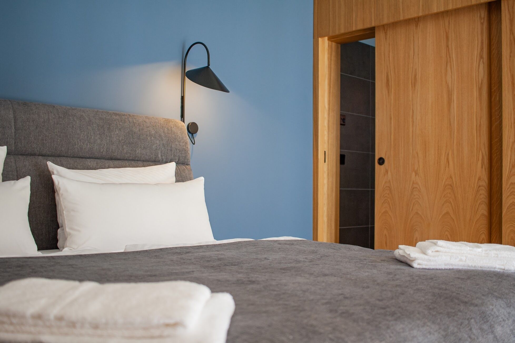 A bedroom with a grey headboard, white pillows, a grey bedspread, a black wall-mounted lamp, and a wooden door.