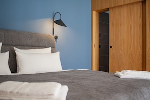 A bedroom with a grey headboard, white pillows, a grey bedspread, a black wall-mounted lamp, and a wooden door. - Project JUNE (Jueterbog)
