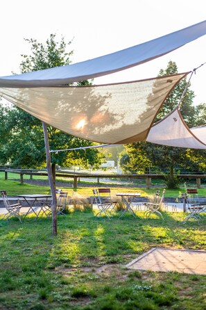 A shaded outdoor seating area with tables and chairs, surrounded by grass and trees. - Project JUNE (Jueterbog)