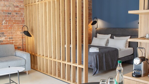 A modern hotel room with a brick wall, a grey sofa, a wooden partition, a bed with grey bedding, a small table with a bottle and glasses, and a coffee machine. - Project JUNE (Jueterbog)