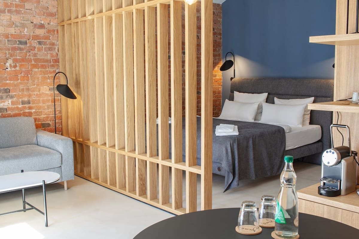 A modern hotel room with a brick wall, a grey sofa, a wooden partition, a bed with grey bedding, a small table with a bottle and glasses, and a coffee machine.
