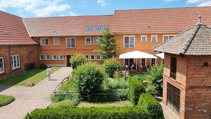 A courtyard with a brick building, a paved walkway, and a small garden area with a table and chairs under a white umbrella. - Project JUNE (Jueterbog)