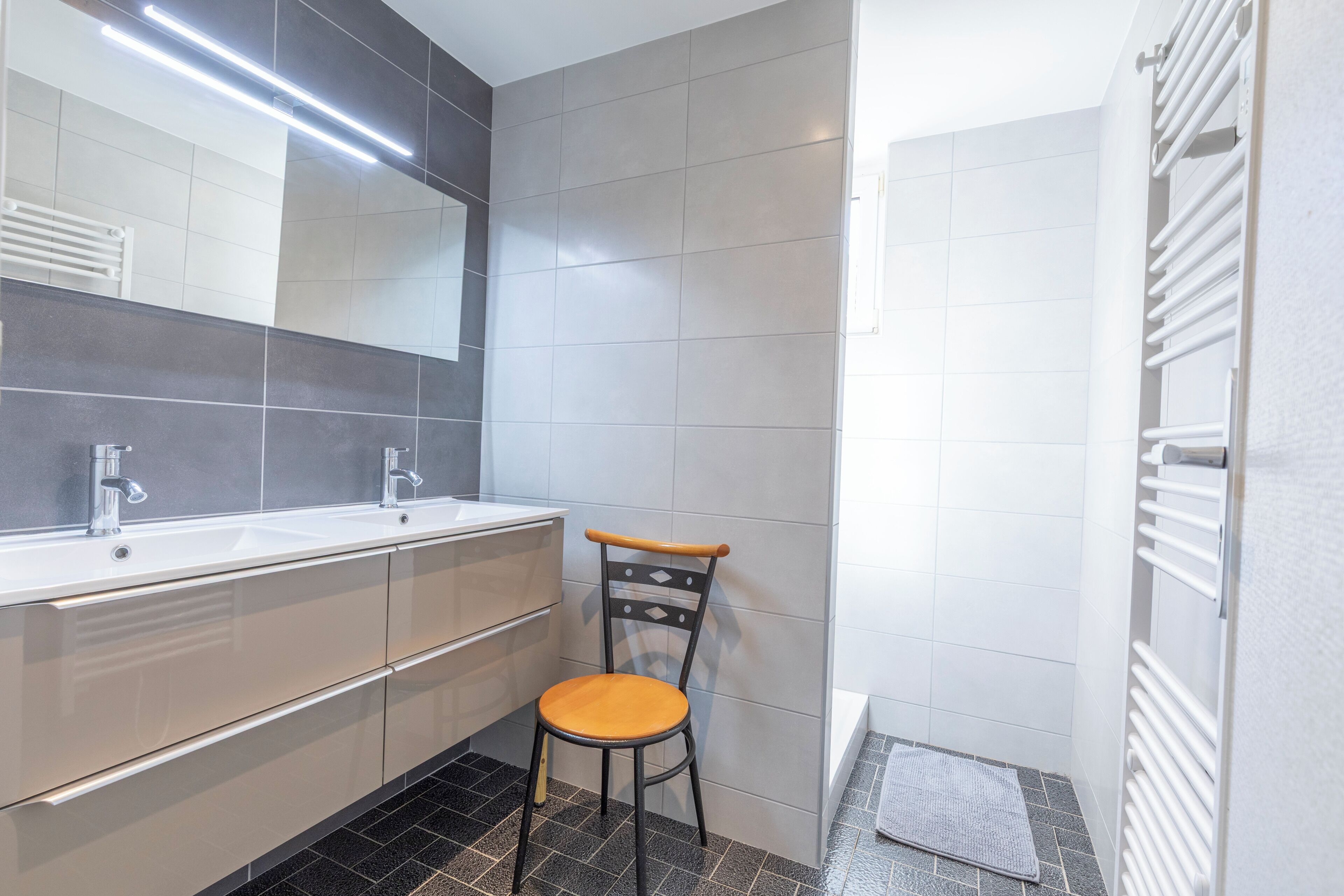 Un baño moderno con tocador de doble lavabo, una silla de madera y un perchero para toallas.