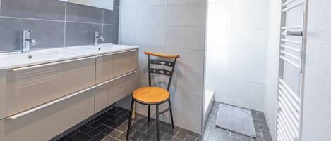Un baño moderno con tocador de doble lavabo, una silla de madera y un perchero para toallas.