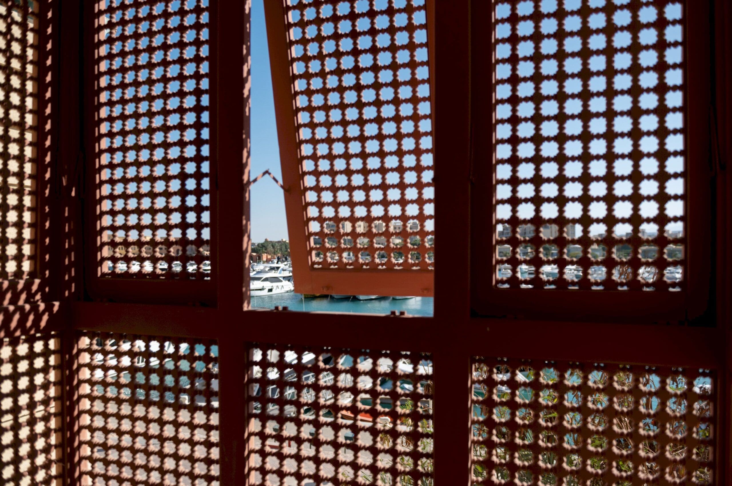 Una ventana con un diseño geométrico, a través de la cual se ve un puerto deportivo con barcos.