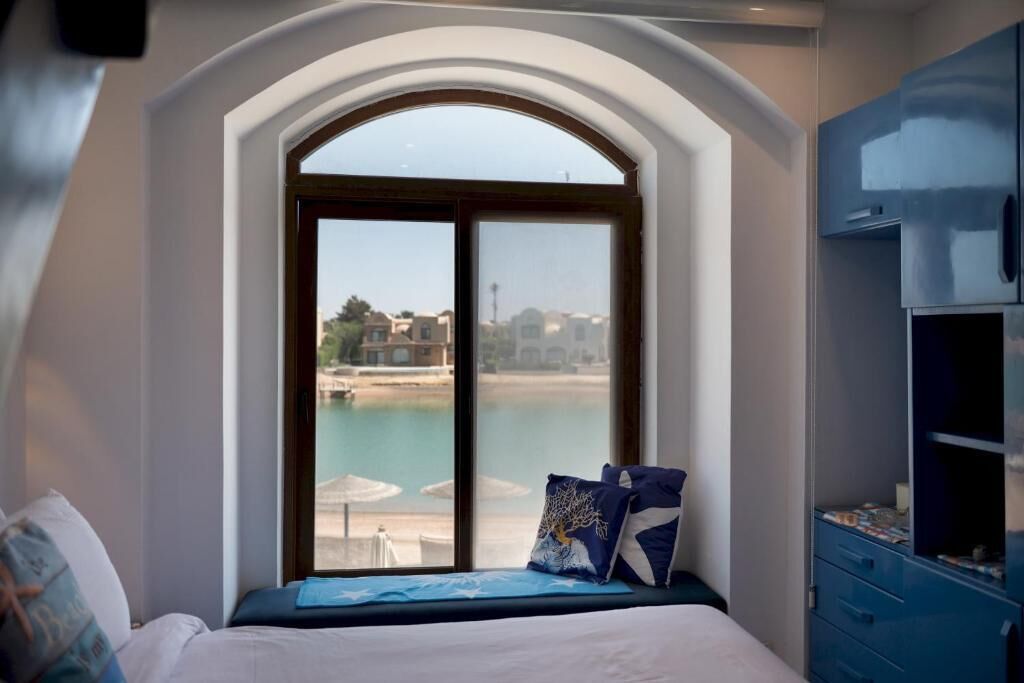 A bedroom with a large arched window overlooking a body of water, a bed with a blue bedspread, and a blue wardrobe.