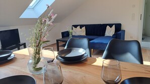 Ein Essbereich mit einem Holztisch, schwarzen Stühlen, einer Blumenvase und einem blauen Sofa im Hintergrund.
