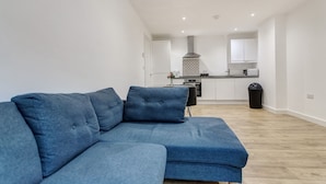 A modern living room with a blue sectional sofa, a kitchen in the background, and a visible trash bin. - 1 Bed, Sleeps 3, Free Parking, Wifi (Slough)