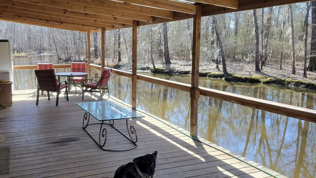 A covered deck with a glass table and chairs, overlooking a calm body of water and a wooded area.