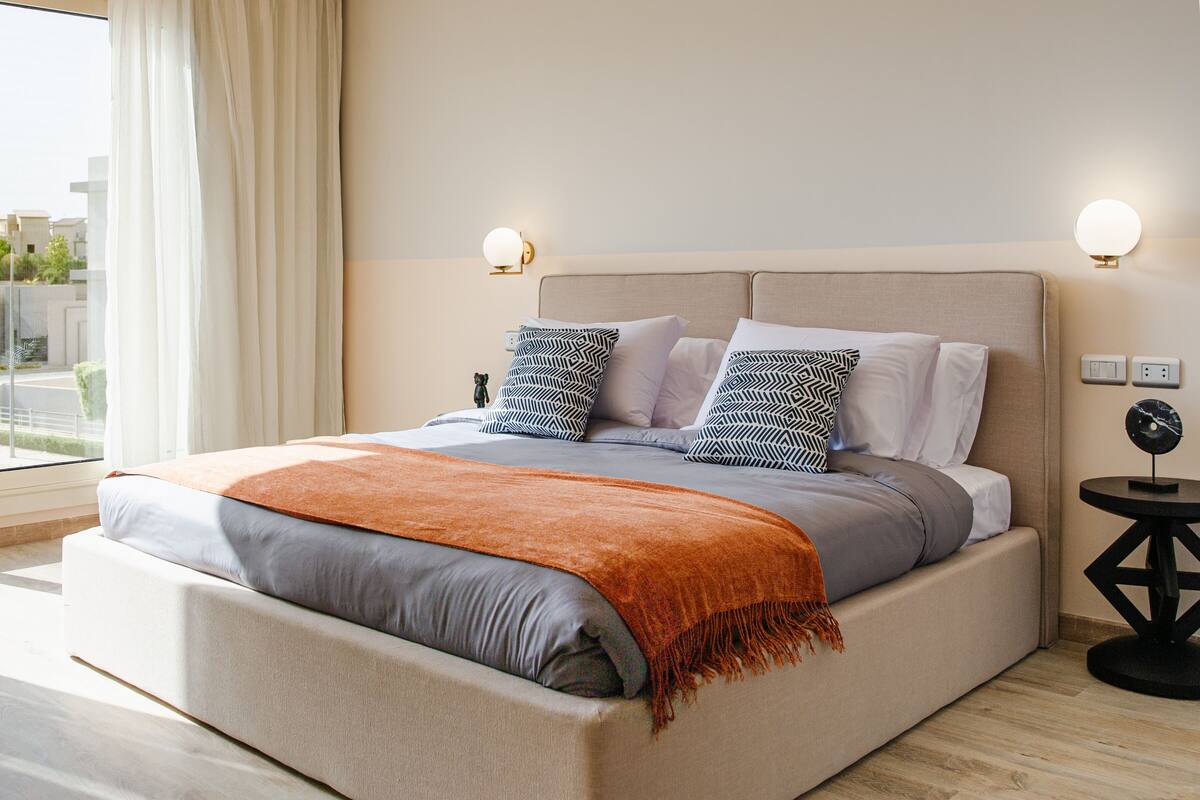 A modern bedroom with a large bed, two bedside lamps, and a view of the city through the window.
