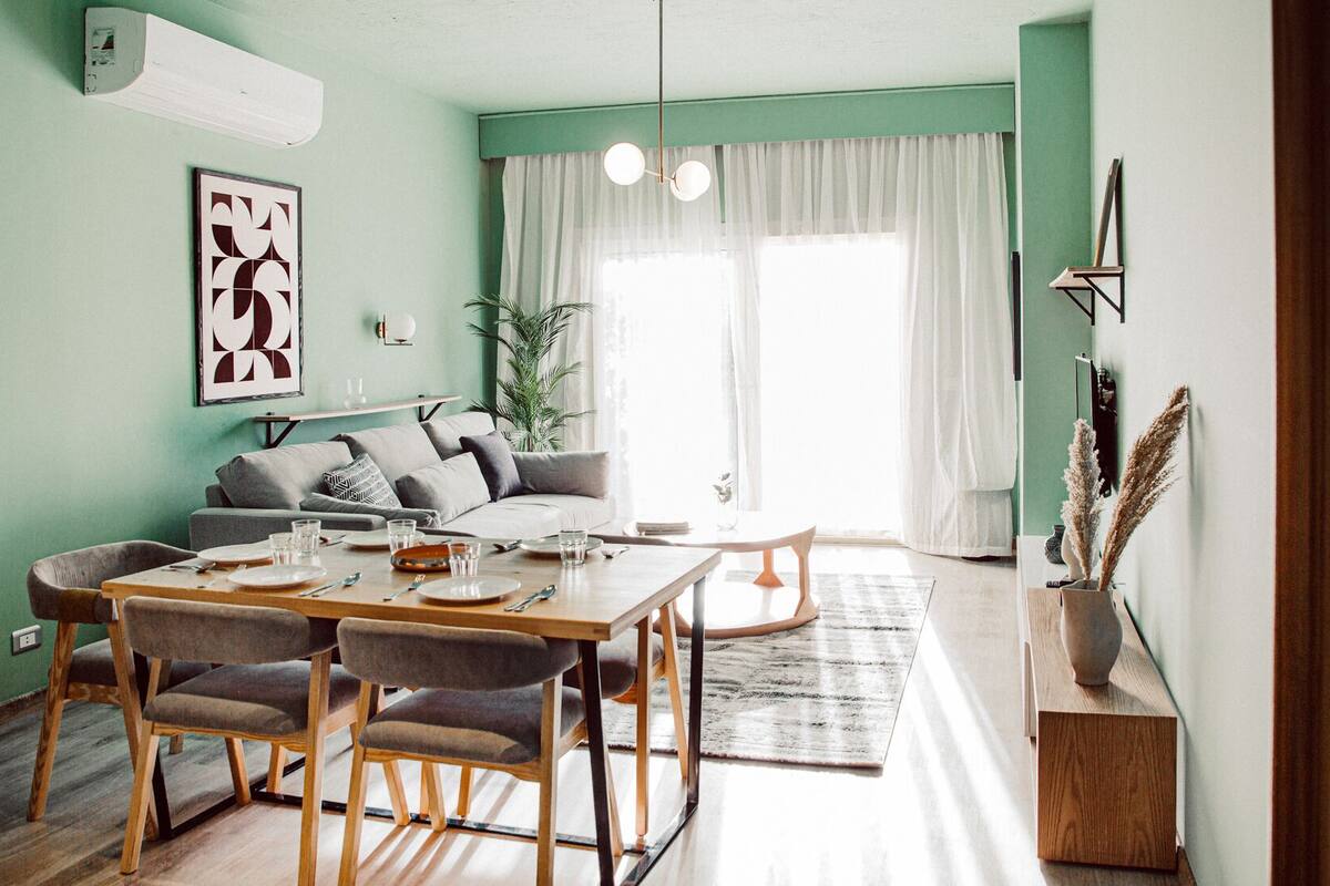 A modern living room with a dining area, a sofa, a coffee table, and a large window with sheer curtains.