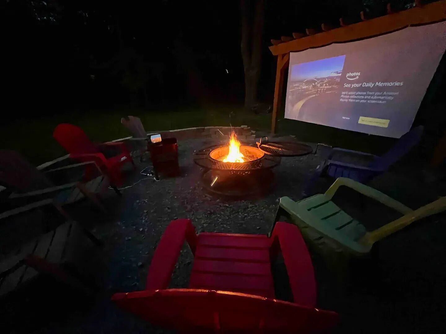 Un espace extérieur accueillant avec un foyer, un écran de projection diffusant un diaporama, et des chaises de jardin rouges et vertes.